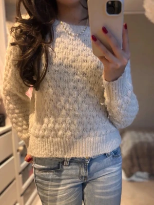 Beige stickad tröja med struktur - Mysig beige stickad tröja med rund hals och långärmade ärmar. Tröjan har ett snyggt strukturerat mönster och ribbade muddar vid ärmslut och nederkant. Perfekt att matcha med jeans för en chill look.