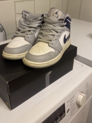 Nike Air Jordan 1 Mid Wolf Grey 38 - Nike Air Jordan 1 Mid sneakers i färgerna vit, grå och marinblå. Klassisk high-top siluett med snörning och rund tå. Swoosh-logga i marinblått på sidan och perforerad tå för extra ventilation. Tillverkade i syntet och skinn för en schysst look.