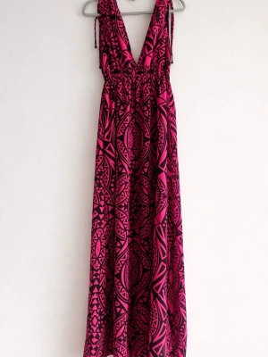 Långklänning med rosa och svart mönster - Snygg långklänning med djup v-ringning och breda axelband som knyts. Klänningen har ett grafiskt mönster i rosa och svart och är lätt och luftig, perfekt för varma dagar. Midjan är smockad för en bekväm passform och tyget faller fint längs kroppen.