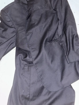 Svart kavaj i ull/polyester, strl 54 - Snygg svart kavaj i klassisk modell, tillverkad av 70% ull och 30% polyester. Kavajen har enkla fickor, knappar framtill och en stilren look som passar perfekt till kostymbyxor. Superfin 140's kvalitet på tyget för en lyxig känsla.