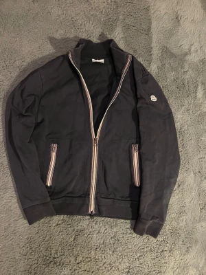 Moncler ziptröja - Mörkblå ziptröja från Moncler med ribbad krage och muddar. Tröjan har dragkedja framtill och två fickor med randiga dragkedjor. Moncler-logga på vänster ärm. Storleken är L men sitter mer som M. Priset går att diskutera.