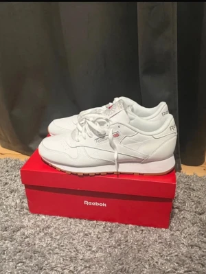 Vita Reebok Classic sneakers - Säljer ett par fräscha Reebok Classic sneakers i helvitt med röda och grå detaljer på tungan. Skorna har snörning, rund tå och platt sula. Materialet är skinn och syntet, vilket ger en clean och stilren look. Perfekta för dig som gillar retrovibbar och enkel design.