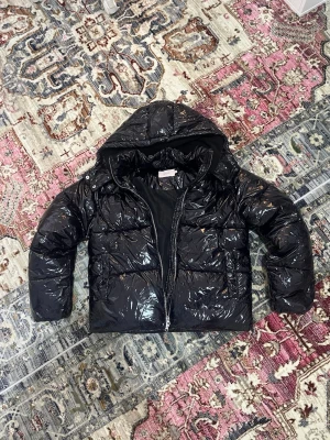Svart glansig pufferjacka från Nelly - Säljer en svart, glansig pufferjacka från Nelly i storlek M. sopass oversized look, med huva och dragkedja framtill. Yttermaterialet är 100% polyamid och fodret är återvunnen polyester. Perfekt för kalla dagar när du vill ha en cool och trendig vibe.