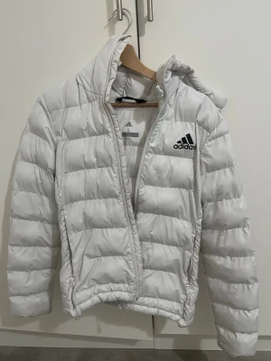 Vit pufferjacka från Adidas - Säljer en vit pufferjacka från Adidas i storlek S. Jackan har huva, dragkedja framtill och Adidas-logga på bröstet. Den är vadderad och har en sportig look som passar perfekt till kyliga dagar. Materialet är syntet och jackan har långa ärmar.