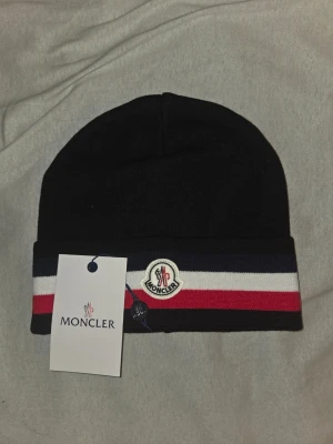 Svart Moncler mössa med ränder - Snygg svart mössa från Moncler med klassiska ränder i marinblått, vitt och rött samt Moncler-logga framtill. Mössan är ribbstickad och har en uppvikt kant. Perfekt accessoar för att lyfta din vinterstil.