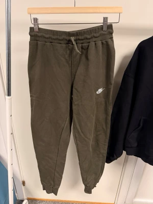 Olivgröna mjukisbyxor från Nike&hoddie svart - Säljer ett par olivgröna mjukisbyxor från Nike med vit logga på benet. Byxorna har resår i midjan med snörning och muddar vid bensluten för en skön och avslappnad passform. Perfekta för chill eller träning.