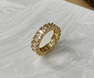 Guldfärgad ring med klara stenar - En chunky guldfärgad ring med gnistrande klara stenar runt hela bandet. Ringen har en lyxig känsla med sina stora, facetterade stenar som fångar ljuset snyggt. Perfekt för dig som vill sticka ut med ett statement-smycke.