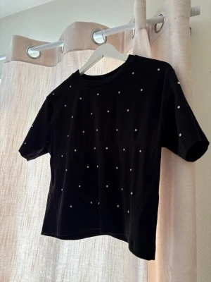 Svart t-shirt med pärldetaljer - Snygg svart t-shirt med korta ärmar och rund hals. T-shirten är dekorerad med små vita pärlor över hela framsidan och ärmarna för en extra cool touch. Tillverkad i mjuk bomull och har en rak, något croppad passform. Använd 1 gång 