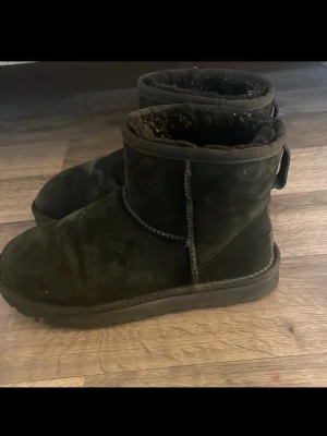 Svarta UGG boots i mocka - Mysiga svarta UGG boots i klassisk modell med rund tå och platt sula. Utsidan är i mjuk mocka och insidan är fodrad med fluffigt material för extra värme. Perfekta för kalla dagar och har en enkel, stilren look. Använda en vinter