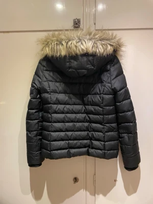 Svart pufferjacka från Tommy Hilfiger - Säljer en svart pufferjacka från Tommy Hilfiger med avtagbar huva och päls. Jackan har dragkedja framtill, två fickor och diskreta loggor på bröstet och ärmen. Perfekt för kalla dagar och riktigt snygg till alla outfits.