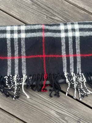 Burberry halsduk - Jovlars fet svart halsduk i mycket fint skick, det finns inga riktiga defekter och halsduken är mycket len och mysig, däremot som man kan se på 2e och 3e bilden så är den vita färgen lite utsmetad men detta är inget som gör halsduken mindre fin utan jag skulle säga motsatsen, skriv vid frågor etc, den är givetvis äkta
