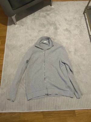 Ljusgrå kofta från Selected Homme - Snygg ljusgrå kofta från Selected Homme med dragkedja framtill och hög krage. Tröjan är stickad i mjukt material och har ribbade muddar vid krage, ärmslut och nederkant. Perfekt för lager-på-lager under kyliga dagar.