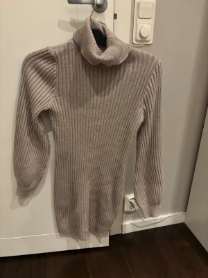Beige ribbad klänning - Säljer en beige ribbad poloklänning med lång ärm och hög krage. Tröjan är stickad och har en slim passform som sitter snyggt på kroppen. Perfekt för att hålla sig varm under kyliga dagar.