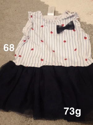 Randig babyklänning med rosett - Supersöt babyklänning med vit och blårandig överdel, små röda nyckelpigor och en mörkblå rosett vid halsen. Kjolen är i mörkblått tylltyg som ger extra volym. Ärmlös modell med volangdetaljer på axlarna. Perfekt för sommarens alla tillfällen!🌸 Fint använt skick. Storlek 68