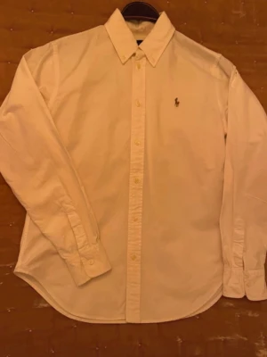 Vit skjorta från Ralph Lauren - Klassisk vit skjorta från Ralph Lauren med button-down krage och broderad logga på bröstet. Skjortan har lång ärm, knappar framtill och är tillverkad i mjuk bomull. Perfekt för en clean och stilren look.