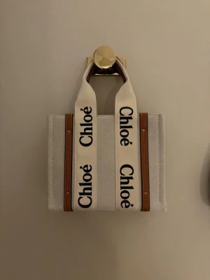  beige axelväska i canvas - Säljer en snygg beige axelväska från Vietnam med breda handtag i canvas med logga. Väskan har bruna skinn-detaljer längs sidorna och är i en stilren, boxig form. Den är inte äkta.