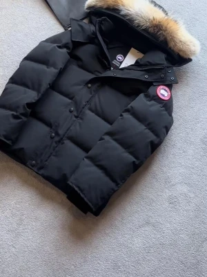 Svart pufferjacka från Canada Goose - Svart pufferjacka från Canada Goose med ikonisk patch på ärmen och fluffig päls på huvan. Jackan har knappar och dragkedja framtill, samt en riktigt schysst dunfyllning som håller dig varm. Perfekt för kalla vinterdagar och har en clean, oversized vibe.