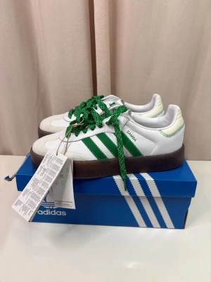 Adidas Samba sneakers vit/grön - Adidas Samba sneakers i vitt skinn med gröna detaljer och gröna snören. Klassiska tre ränder på sidan och mörkbrun gummisula. Låg modell med rund tå och retrovibe. Perfekta för dig som gillar streetstyle och tidlös design.