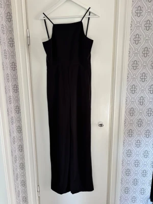 Svart byxdress med smala axelband - Säljer en svart byxdress med smala justerbara axelband och rak halsringning. Byxdressen har vida ben och en diskret dragkedja i ryggen. Materialet känns mjukt och har ett snyggt fall, perfekt för dig som gillar stilrena plagg med enkel design. Sparsamt använd. 