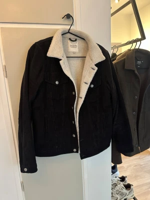 Svart teddyjacka från Just Junkies - Svart teddyjacka från Just Junkies i manchestertyg med vit fluffig insida och krage. Klassisk rak passform, knappar framtill och två bröstfickor med lock. Perfekt för kalla dagar och riktigt snygg till jeans.