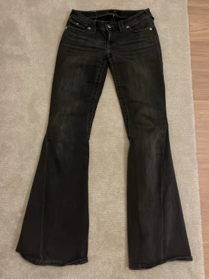 Svarta bootcut jeans True Religion - Säljer ett par svarta jeans från True Religion med bootcut passform. Byxorna har klassiska fem fickor, silverfärgade knappar och detaljerade bakfickor med lock. Materialet är jeans och färgen är mörk svart med lite urtvättad look. Lite trasiga längst ner pga för långa men de går att sy upp ifall de stör!