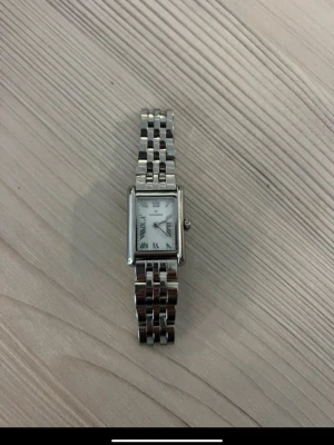Mockberg klocka  - Elegant and timeless Mockberg watch in silver, perfect for everyday wear or special occasions. Minimalist rectangular design that never goes out of style.  🔹 Condition: Very good condition – well cared for 🔹 Original price: 1800 SEK 🔹 Color: Silver 🔹 Material: Stainless steel 🔹 Details: Roman numerals, classic and feminine look  📦 Comes with original box + extra links (easy to adjust the size)  A perfect gift for yourself or someone special 🤍 Selling only because it’s not getting enough use.  