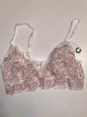Blommig spetsbralette från Cubus - Bralette från Cubus i triangelformad modell med vit och ljusrosa blommig spets. Justerbara axelband och hakknäppning bak. Oanvänd, nypris 200.