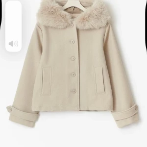 Beige kappa med fuskpäls - Snygg beige kappa med stor, fluffig fuskpäls runt huvan. Jackan har raka fickor, knappar framtill och dekorativa band med knapp vid ärmsluten. Perfekt för dig som vill ha en stilren och mysig look under kyliga dagar.