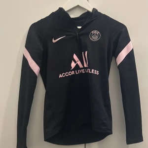 Svart långärmad PSG fotbollströja - Snygg svart långärmad fotbollströja från Nike med Paris Saint-Germain-logga på bröstet och rosa detaljer på ärmarna samt tryck framtill. Tröjan har halv dragkedja vid halsen och är gjord i ett lätt, sportigt material som passar perfekt för träning.