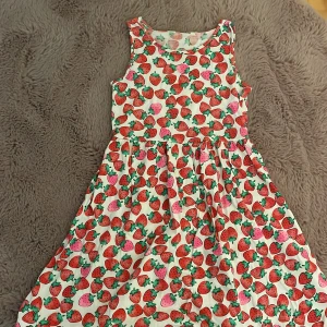 Jordgubbsmönstrad klänning från H&M - Superfin ärmlös klänning från H&M med ett färgglatt jordgubbsmönster på vit botten. Klänningen har rund halsringning och ett lätt utställt kjolparti.