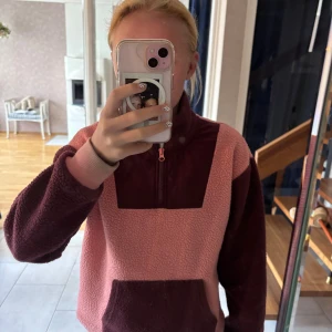 Rosa och vinröd fleece hoodie - Mysig hoodie i teddyfleece med färgblock i rosa och vinrött. Tröjan har half zip framtill, stor magficka och ribbade muddar. Perfekt för kyliga dagar och riktigt skön att dra på sig när man vill vara bekväm.