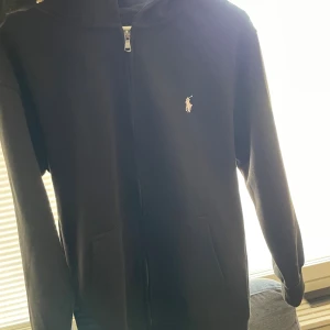 Svart hoodie från Ralph Lauren - Svart hoodie från Ralph Lauren med dragkedja framtill och klassisk vit logga på bröstet. Tröjan har huva, fickor och långa ärmar. Perfekt för chill dagar eller när du vill ha en clean och enkel stil.