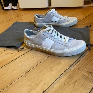 Tom Ford skor  - Snygga vita sneakers från Tom Ford med klassisk låg profil och rund tå. Skorna har en mix av mocka och skinn, vita snören och en vit platt sula. Tom Ford-logga på hälen och stilrena detaljer på sidorna. Perfekt för dig som gillar minimalistisk och lyxig stil.