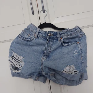 Blåa ripped jeansshorts från Ginatricot - ⚠️ Skriv till mig innan köp!⚠️Snygga blå jeansshorts från Ginatricot med hög midja och slitna detaljer framtill. Klassisk femficksmodell, knappar i silver och uppvikta benslut. Perfekta för sommaren och ger en cool, avslappnad vibe.