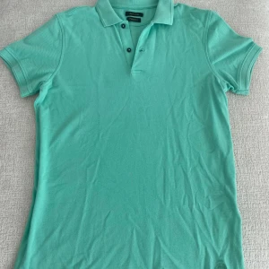 Turkos pikétröja från Massimo Dutti - Snygg turkos pikétröja från Massimo Dutti med klassisk krage och tre knappar framtill. Tröjan har korta ärmar och är tillverkad i mjuk bomull, perfekt för varma dagar. Enkel och stilren design som passar till det mesta.