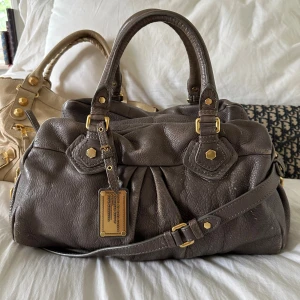 Brun grå Q Classic Boston Bag - Perfekta skolväskan! Rymlig mörkgrå Q Classic Boston bag från Marc by Marc Jacobs i äkta läder med guldfärgade detaljer och dubbla handtag. Långa bandet medföljer. Klassisk och tidlös modell som rymmer allt du behöver och passar perfekt både till vardag och resa. Praktiska fack och dragkedja gör den enkel att organisera. Välbevarad vintage-modell, men slitage finns. Se alla bilder.  Mått 40x22x15 (undersida). Kan hämtas i Nacka eller skickas. Skriv privat för fler bilder 🤍