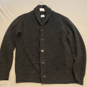 Mörkgrå stickad kofta från Zara - Säljer en mörkgrå, ribbstickad kofta från Zara med snygg sjalkrage och stora knappar framtill. Koftan har lång ärm och är perfekt för lager-på-lager-stil under kyliga dagar. Enkel att matcha och superbekväm.