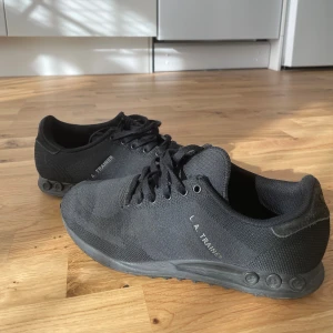 Svarta Adidas LA Trainer sneakers - Säljer ett par helsvarta Adidas LA Trainer sneakers med klassisk design. Skorna har ovandel i textil, snörning och diskreta ränder på sidorna. Sulan har de ikoniska pluggarna och hela skon är svart för en clean look.