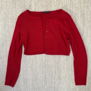 Röd croppad kofta från Brandy Melville - Intressekoll: Snygg röd croppad kofta från Brandy Melville med rund hals och långa ärmar. Koftan är ribbad och har knappar framtill. Perfekt att styla med höga jeans eller kjol för en trendig look.