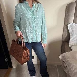 Ljusblå prickig blus med volang - Snygg ljusblå blus med vita prickar och volangdetaljer vid halsen. Långärmad med smock vid ärmslut och knappar framtill. Luftigt material som känns skönt mot huden och ger en chill vibe. Perfekt för dig som gillar söta detaljer och en avslappnad stil.