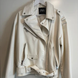Vit skinnjacka från Zara med bälte - Säljer en snygg vit skinnjacka från Zara med coola dragkedjor, fickor och bälte i midjan. Jackan har klassisk bikerstil med bred krage och silverdetaljer. Perfekt för dig som vill sticka ut med en fräsch look.