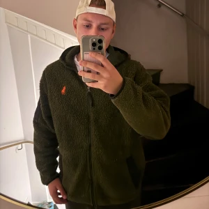 Grön teddyjacka från Polo Ralph Lauren - Mörkgrön teddyjacka från Polo Ralph Lauren med orange logga på bröstet. Jackan har huva, hel dragkedja och två sidofickor. Materialet är mjuk fleece som ger en chill och skön vibe. Perfekt för kalla dagar när du vill vara både bekväm och snygg. Använd en del och har blivit lite noppig 