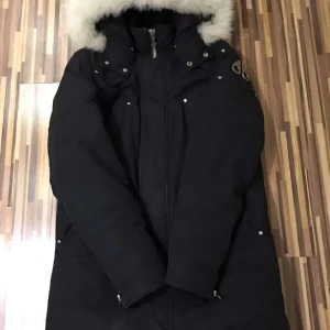 Svart parkasjacka med fuskpäls Moose Knuckles  - Säljer en svart parkasjacka från Moose Knuckles med huva och vit fuskpäls. Jackan har långa ärmar, dragkedja framtill och detaljer i silver. Perfekt för kalla dagar med sin långa modell och snygga design.