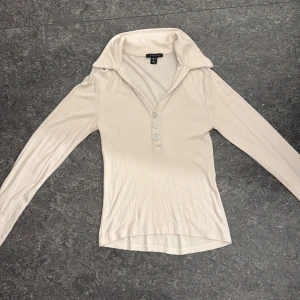 Beige ribbad långärmad topp Amisu - Snygg beige ribbad topp från Amisu med lång ärm och stor krage. Toppen har tre glittriga knappar framtill som ger en extra touch. Perfekt för dig som gillar stilrena och trendiga plagg med lite extra detalj.