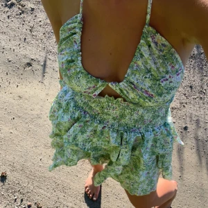 Grön blommig kort sommarklänning - Supersöt kort klänning med grönt och vitt blommönster, volangkjol och smockad midja. Klänningen har v-ringning, tunna axelband och är ärmlös. 