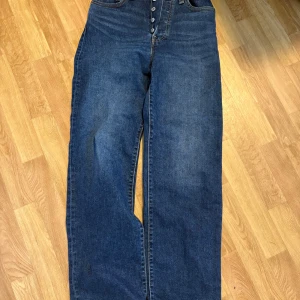 Levi's Ribcage Straight jeans blå 26 - Säljer ett par Levi's Ribcage Straight jeans i klassisk blå denim. Hög midja, raka ben och knäppning med knappar framtill. Snygga bakfickor med Levi's röda etikett. Perfekta för dig som gillar en tidlös och clean look.