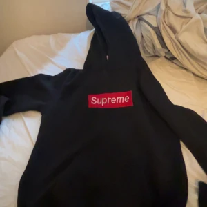 Svart Supreme hoodie med röd logga - Supreme hoodie i svart med den klassiska röda boxloggan framtill. Tröjan har huva och långa ärmar, perfekt för dig som vill ha en streetwear-look. Mjuk och skön, passar till många outfits.