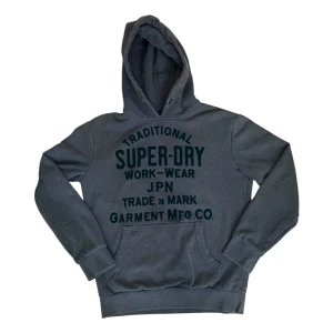 Superdry hoodie  - Säljer en mörkgrå hoodie från Superdry med stor logotyp och text framtill. Tröjan har huva med snörning och en klassisk magficka.