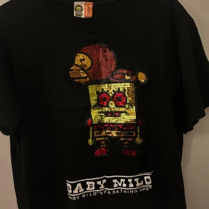 Svart Baby Milo t-shirt med paljetter - Limited edition, Baby Milo x SpongeBob T-shirt (BAPE) Storlek: M Svart t-shirt med unik paljett-design av Baby Milo & SpongeBob. Skimrande detaljer i guld/rött/gult paljetter. Ny skick. ORIGINAL