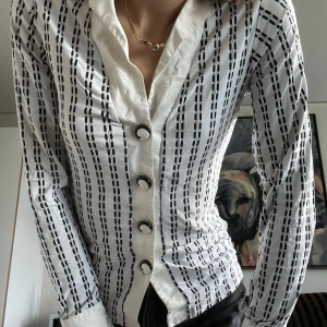 Vit och svart randig skjorta med stora knappar - Säljer en vit skjorta med svarta vertikala ränder och stora, runda knappar framtill. Skjortan har lång ärm, klassisk krage och markerade manschetter. Snygg slim passform och tillverkad i ett lätt bomullstyg. Perfekt för dig som gillar retrovibbar och grafiska mönster.